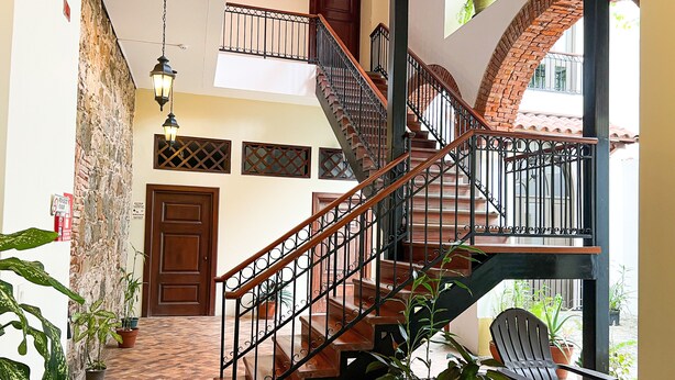 A staircase with wooden steps and black railings, leading to a landing with a door. There are potted plants on the landing and a chair near the door. - Apartment in the Heart of Casco Viejo Panama 11 (Panamá)