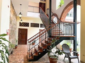 A staircase with wooden steps and black railings, leading to a landing with a door. There are potted plants on the landing and a chair near the door.