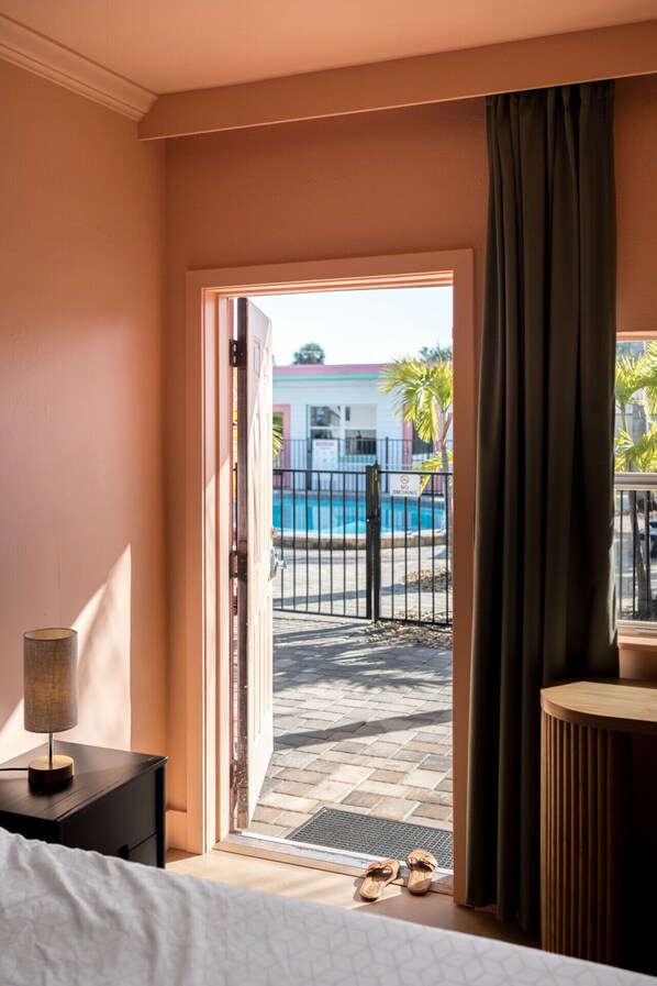 A hotel room with a bed, a nightstand, a lamp, a view of a swimming pool, and a door leading outside.