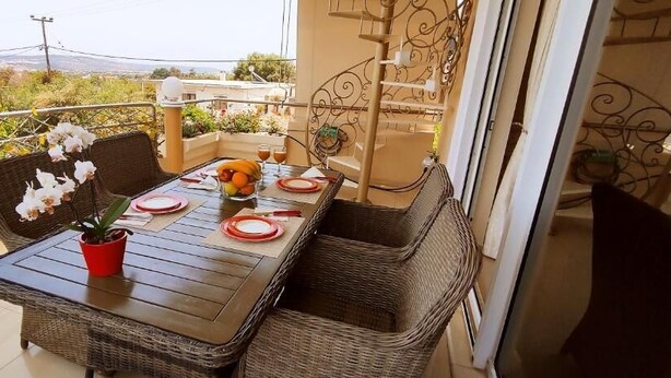 Un balcón con mobiliario de mimbre, una mesa dispuesta con platos, una maceta roja y una planta en maceta.