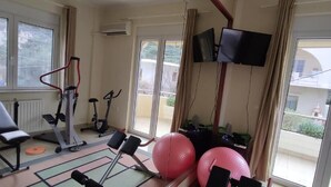 Una habitación con una cinta caminadora, bicicleta estática y pelotas de ejercicios, ventanas amplias y un televisor montado en la pared.