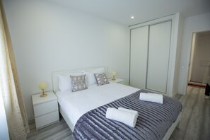 A neatly made bed with a grey blanket and two rolled towels, a bedside table with a lamp, and a wardrobe with sliding doors. - Alecrm D Estepe - Hostel (Almada)
