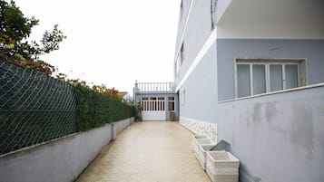 A narrow pathway leading to a building entrance with a tiled floor, a chain-link fence on one side, and a white wall on the other.