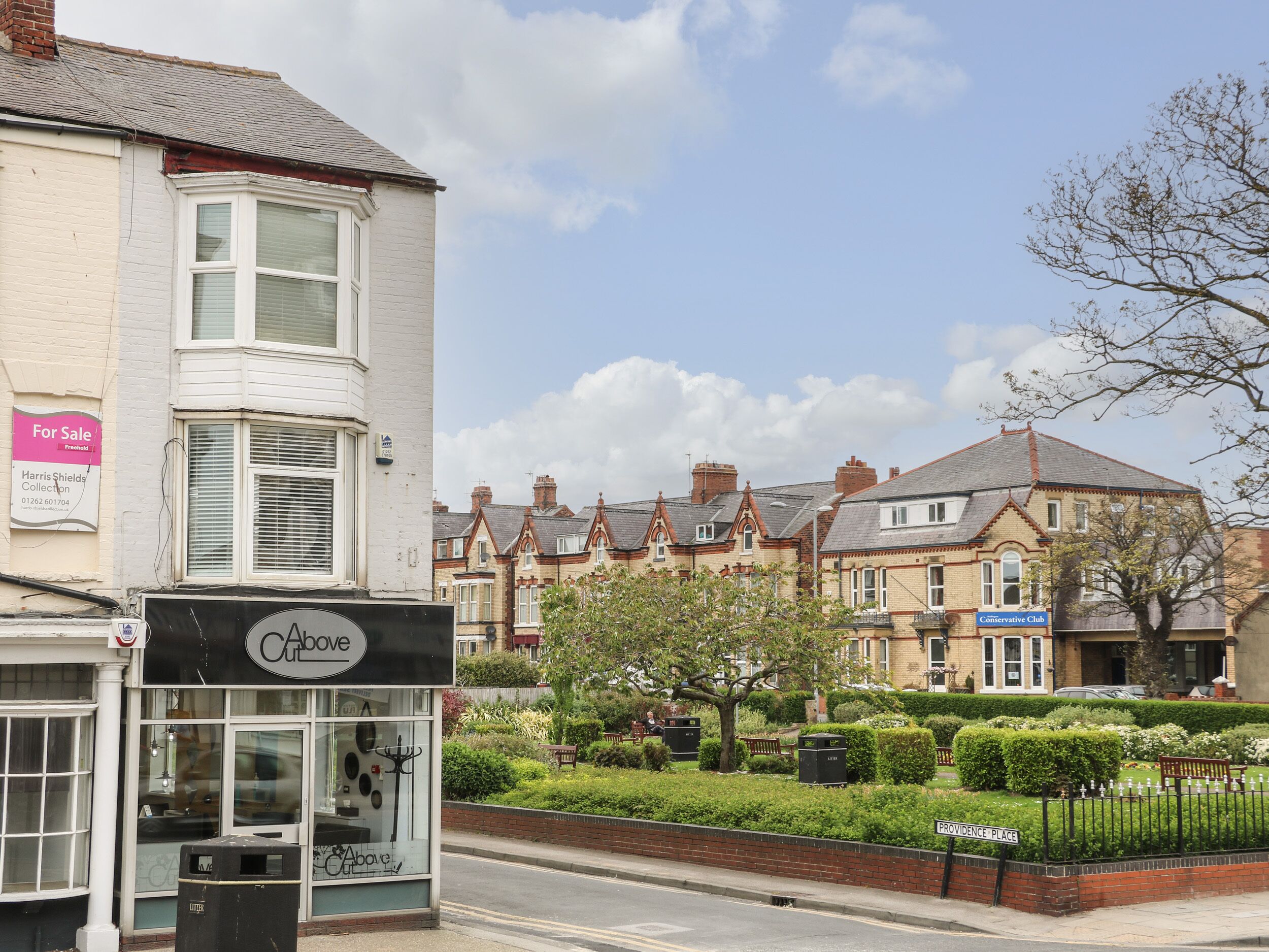 A street view with a cafe named 'Cafe Above' on the left, a 'For Sale' sign, and a 'Conservative Club' sign on the right.