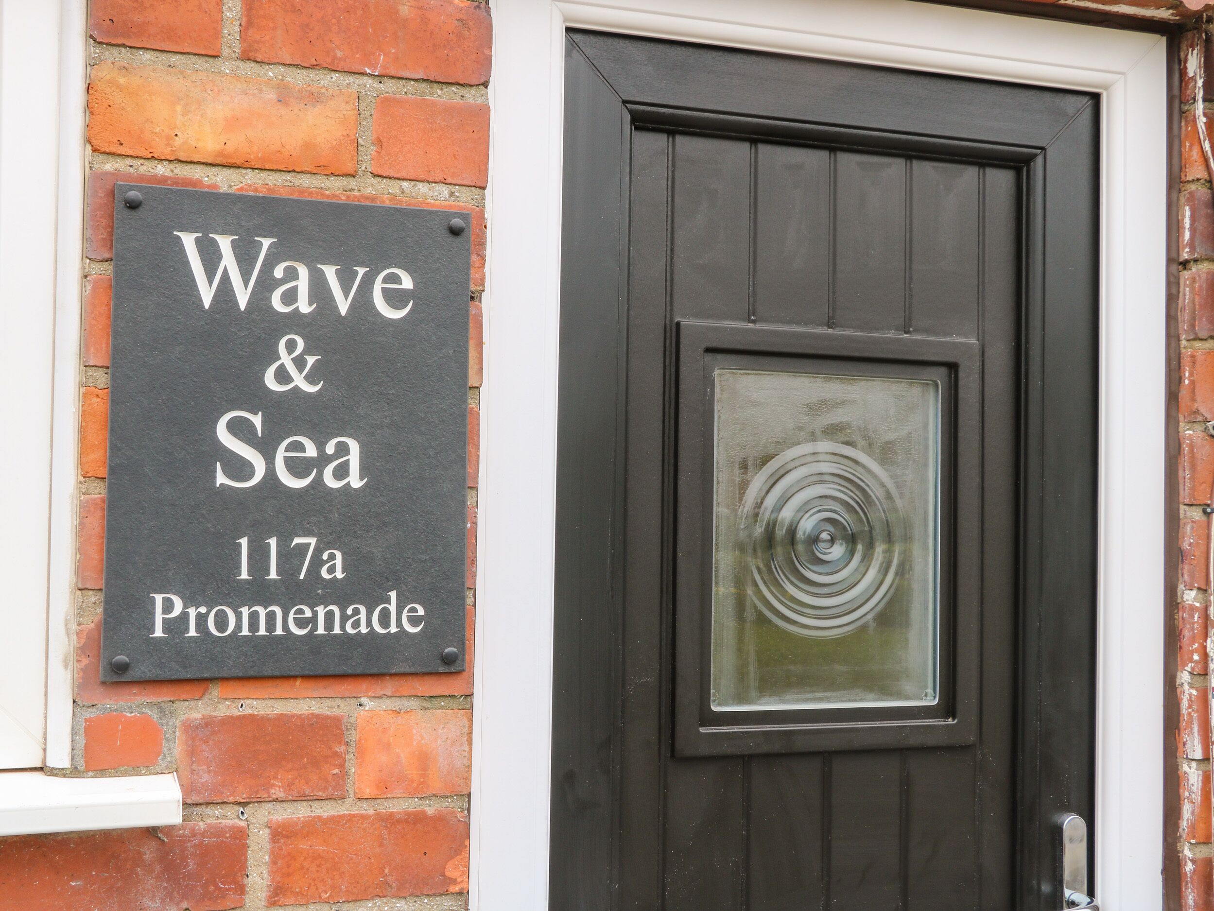 A black sign with white lettering reading "Wave & Sea 117a Promenade" mounted on a brick wall next to a dark wooden door.