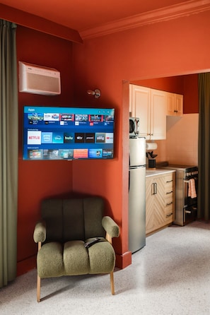 A room with a TV mounted on the wall displaying various streaming apps, a sofa, a kitchen area with cabinets and a refrigerator, and a microwave.