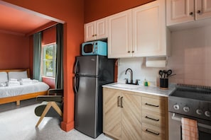 A compact hotel room with a bed, a kitchenette, a refrigerator, a microwave, a sink, and a stove.