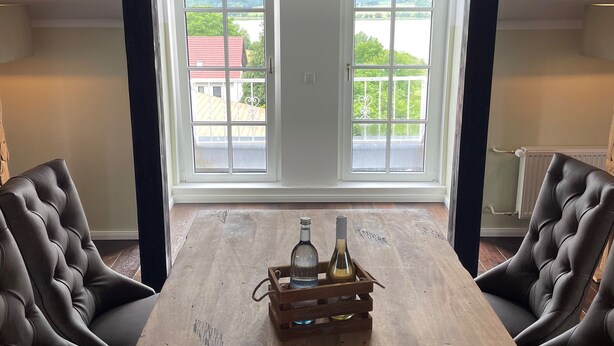 Een kamer met een grote houten tafel, twee leren stoelen en uitzicht op een meer door twee ramen.