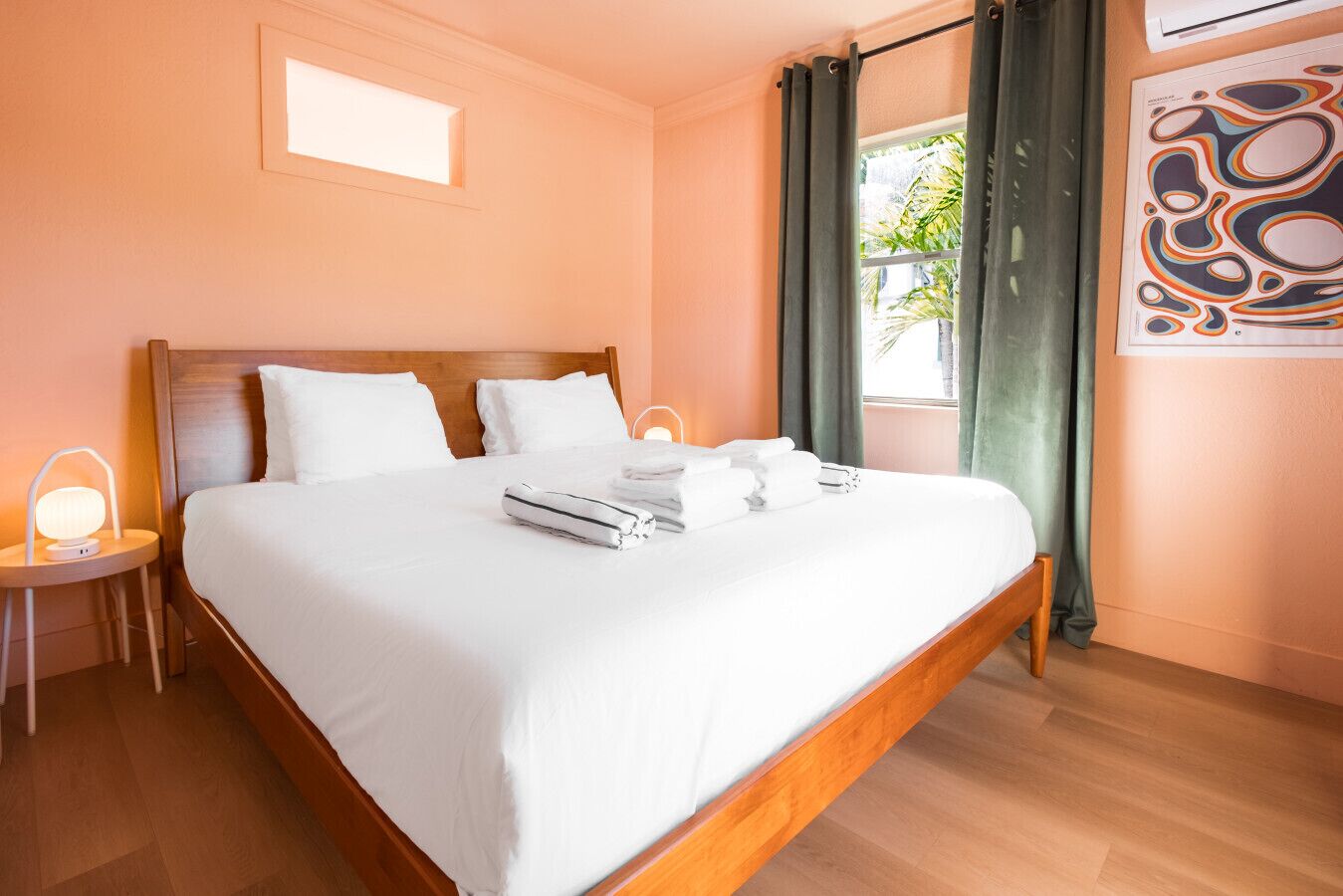 A hotel room with a wooden bed, white linens, a bedside lamp, a window with curtains, and a wall-mounted artwork.