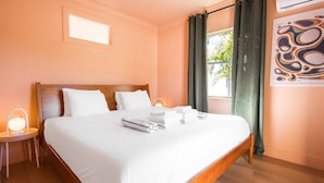 Hotelkamer met een houten bed, witte lakens, een bedlamp, een raam met gordijnen en een kunstwerk aan de muur.