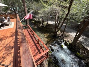 Eine Holzterrasse mit Sitzgelegenheiten, mit Blick auf einen Bach und Bäume.