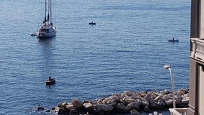 A coastal scene with a sailboat and a small motorboat in the water, a rocky shoreline, and a building with classical architecture. - Di Palma Suite Museum (Napoli)