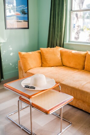 A room with a sofa, a small table with a hat and glasses, and a window with curtains.