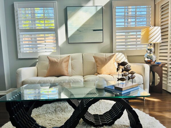 A living room with a glass coffee table, a white sofa with two cushions, a decorative fish sculpture, and a lamp on a wooden side table.