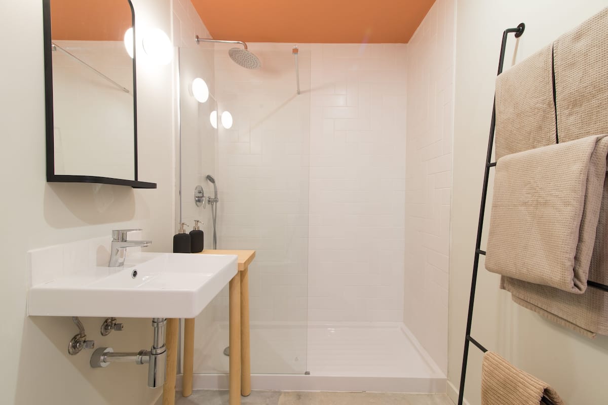 Uma casa de banho moderna com uma sanita branca, espelho e chuveiro. Toalhas pendem de um porta-toalhas e um banquinho de madeira está perto da sanita.