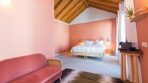 Une chambre avec un plafond en bois, un lit avec une literie blanche, un canapé rouge, une petite table avec un ordinateur portable et une plante posée sur une étagère.