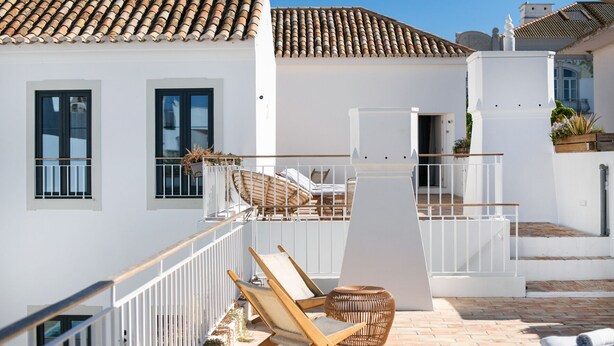 Un bâtiment blanc avec un toit recouvert de tuiles, un balcon doté d’une balustrade blanche et des sièges à l’extérieur.