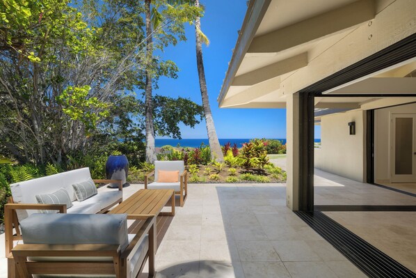 An outdoor patio with a wooden table and chairs, a white sofa, and a view of the ocean. - Expansive Mauna Kea Resort Home, 3 private bdrms and massive kids suite! (Kamuela)