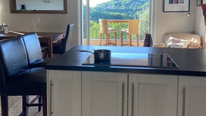 Una cocina con una isla grande, un comedor con mesa y sillas, y vista a un paisaje verde a través de un ventanal amplio.