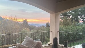 Un balcón acogedor con sofá, cojines y una pequeña mesa, con vistas a un paisaje pintoresco y una puesta de sol.