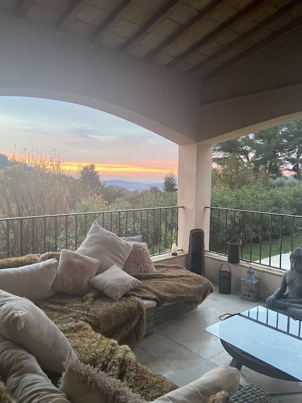 A cozy balcony with a sofa, cushions, and a small table, overlooking a scenic landscape with a sunset view.