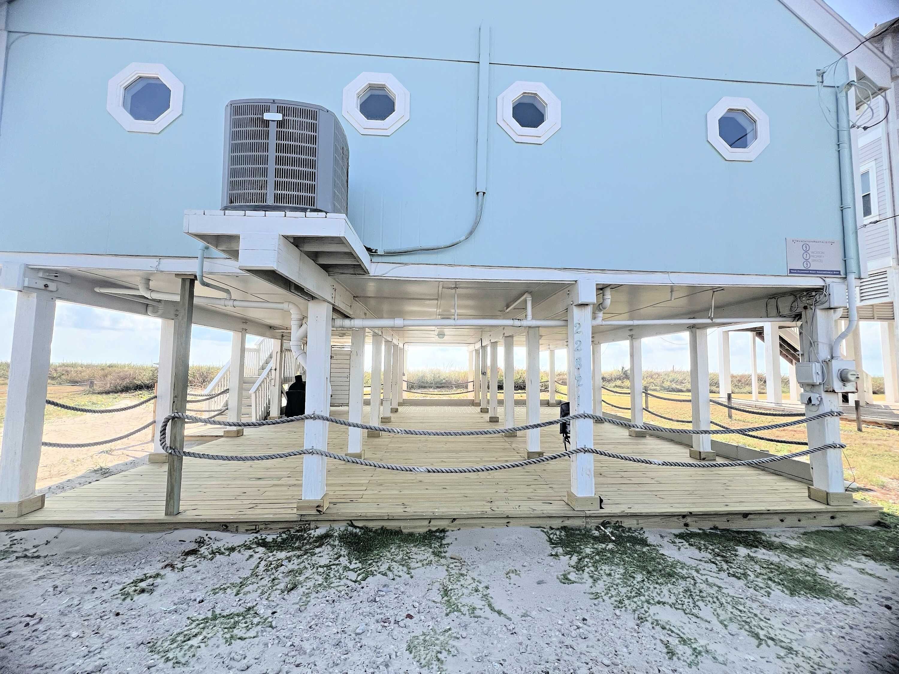 A large, light blue building with multiple windows, a covered deck area, and a sandy ground.