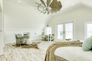 Une pièce spacieuse avec un plafond en pente, un grand lit doté d’une tête de lit en osier, un bureau avec une chaise et une vue sur l’océan grâce à de grandes fenêtres.