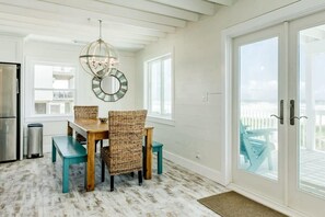 Un coin repas avec une table en bois et des chaises en osier, un miroir suspendu et, derrière une porte vitrée, une vue sur une terrasse extérieure.