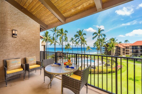 A balcony with wicker furniture, a table with a fruit bowl, and a view of the ocean and palm trees.