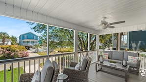 A covered porch with wicker furniture, a ceiling fan, and a view of a blue house and greenery. - Garden City Retreat with Pool, Walk to Beach (Garden City Beach)