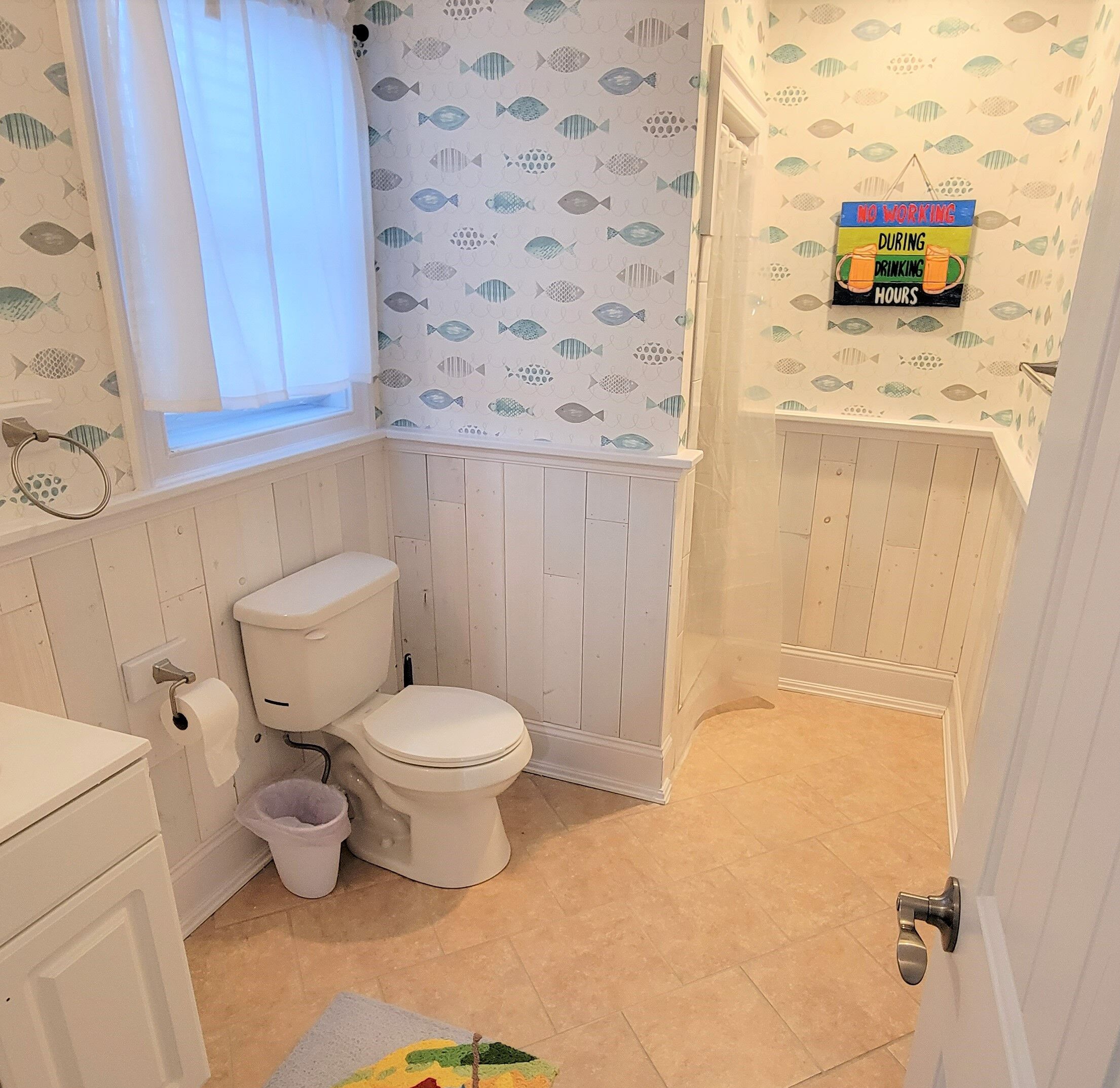 Une salle de bain comprenant des toilettes, un lavabo et un espace douche. Il y a une fenêtre avec un rideau, et les murs sont décorés de motifs représentant des poissons.