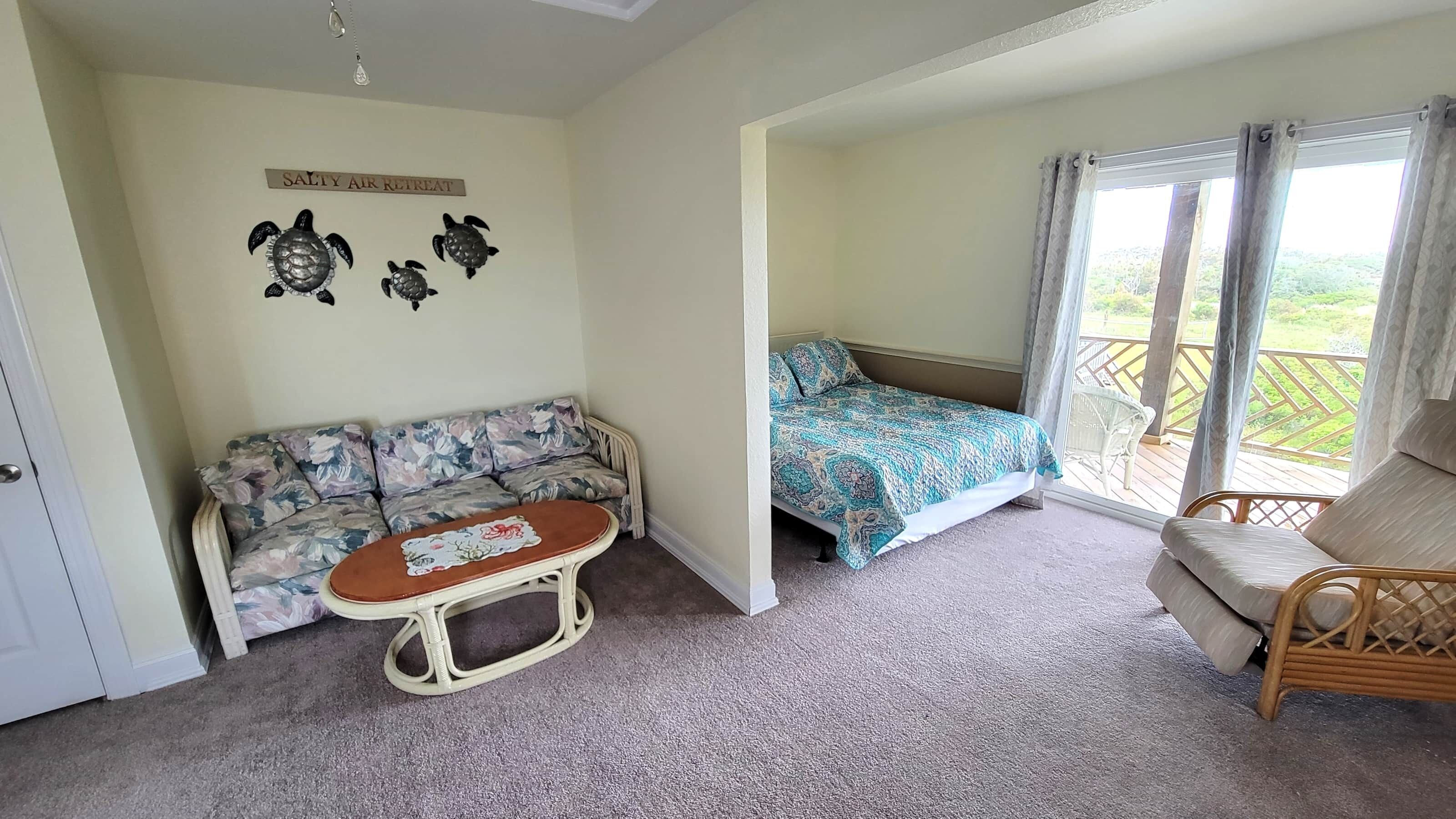 A room with a sofa, a bed with a patterned bedspread, a wicker chair, a small table with a placemat, and a sign on the wall.