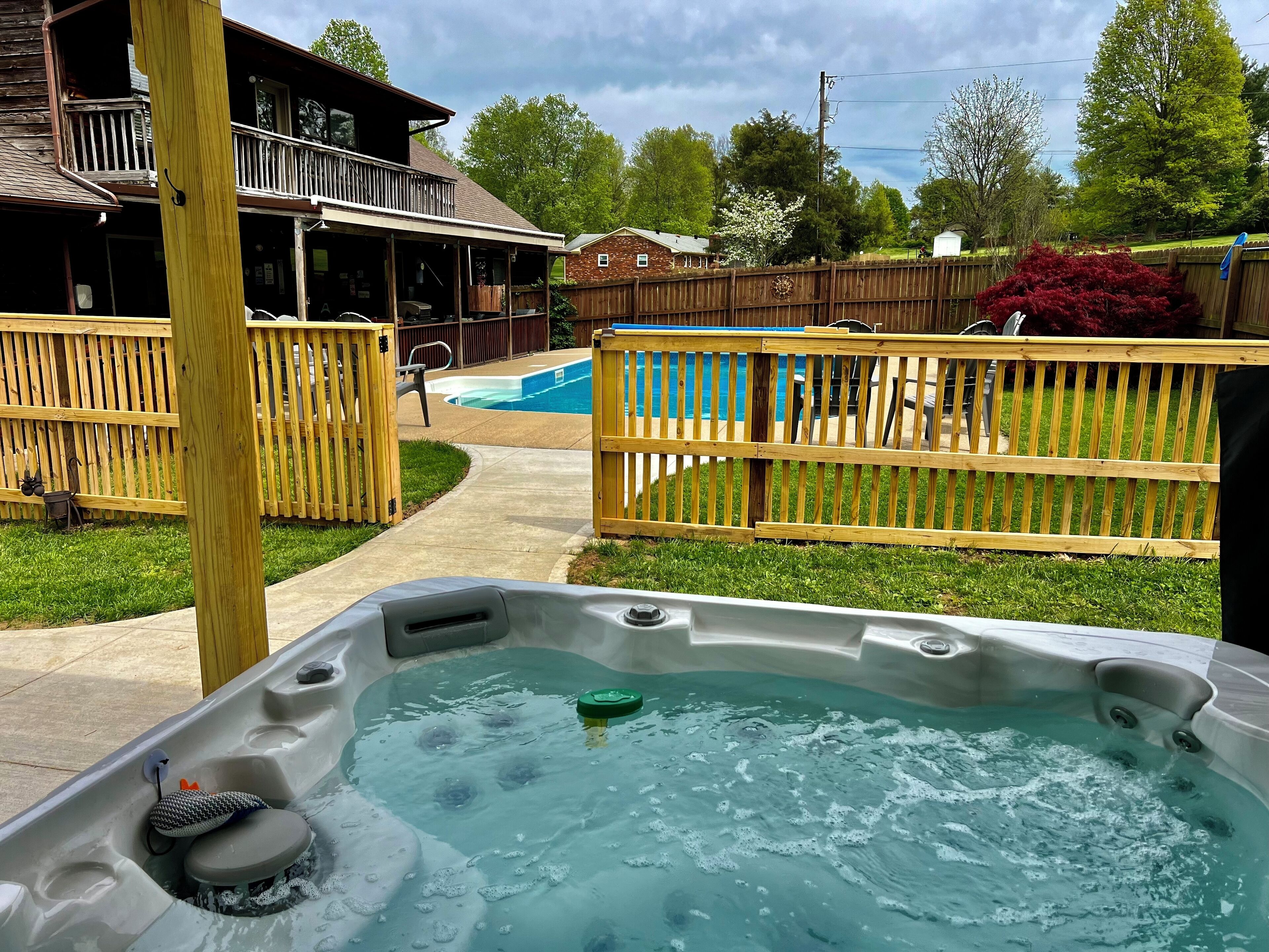 A hot tub in a backyard with a wooden fence, a pool, and a house in the background.