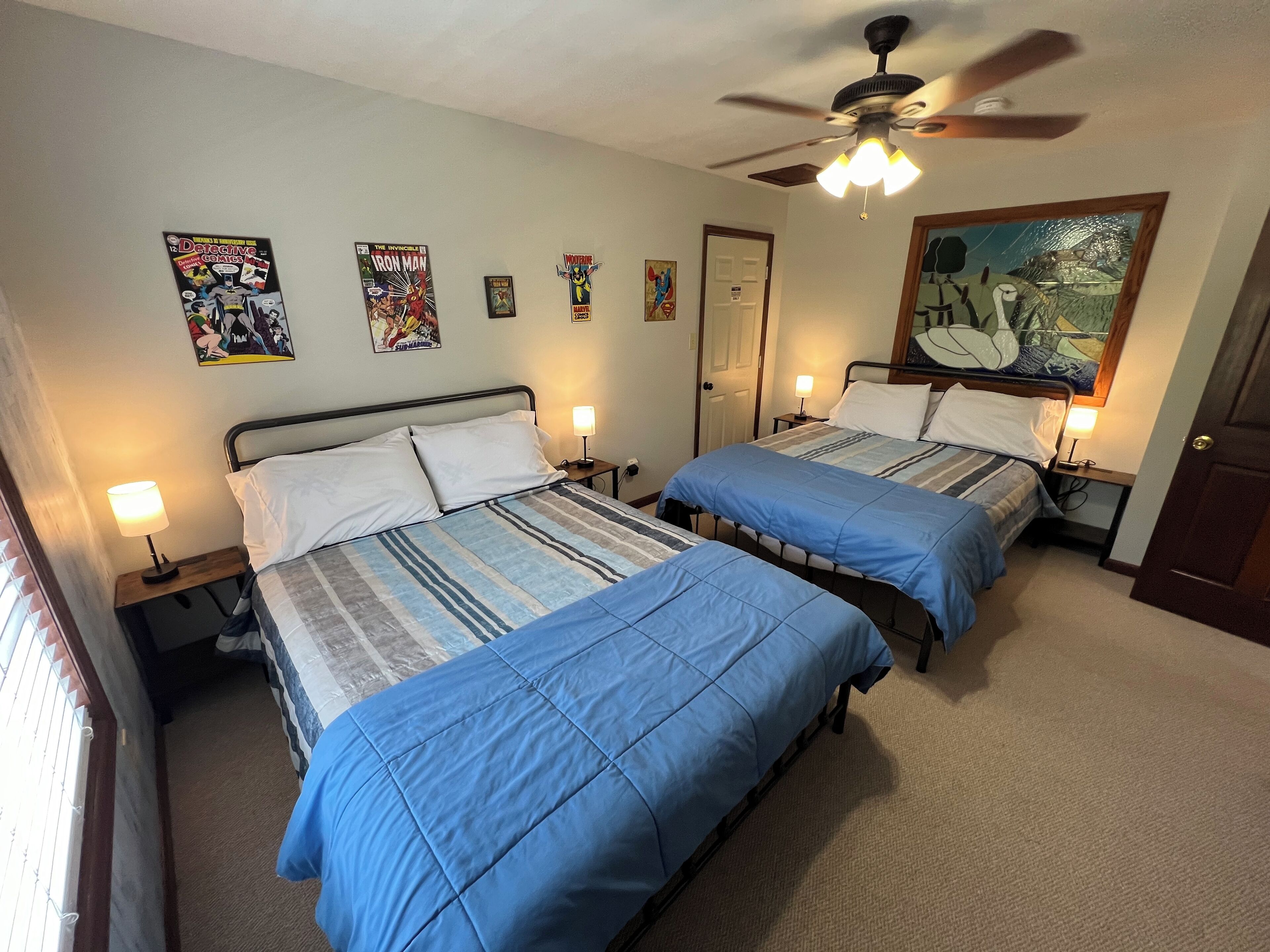 A hotel room with two beds, each with a blue bedspread and white pillows. There are wall-mounted posters, a ceiling fan, and a door with a mirror.