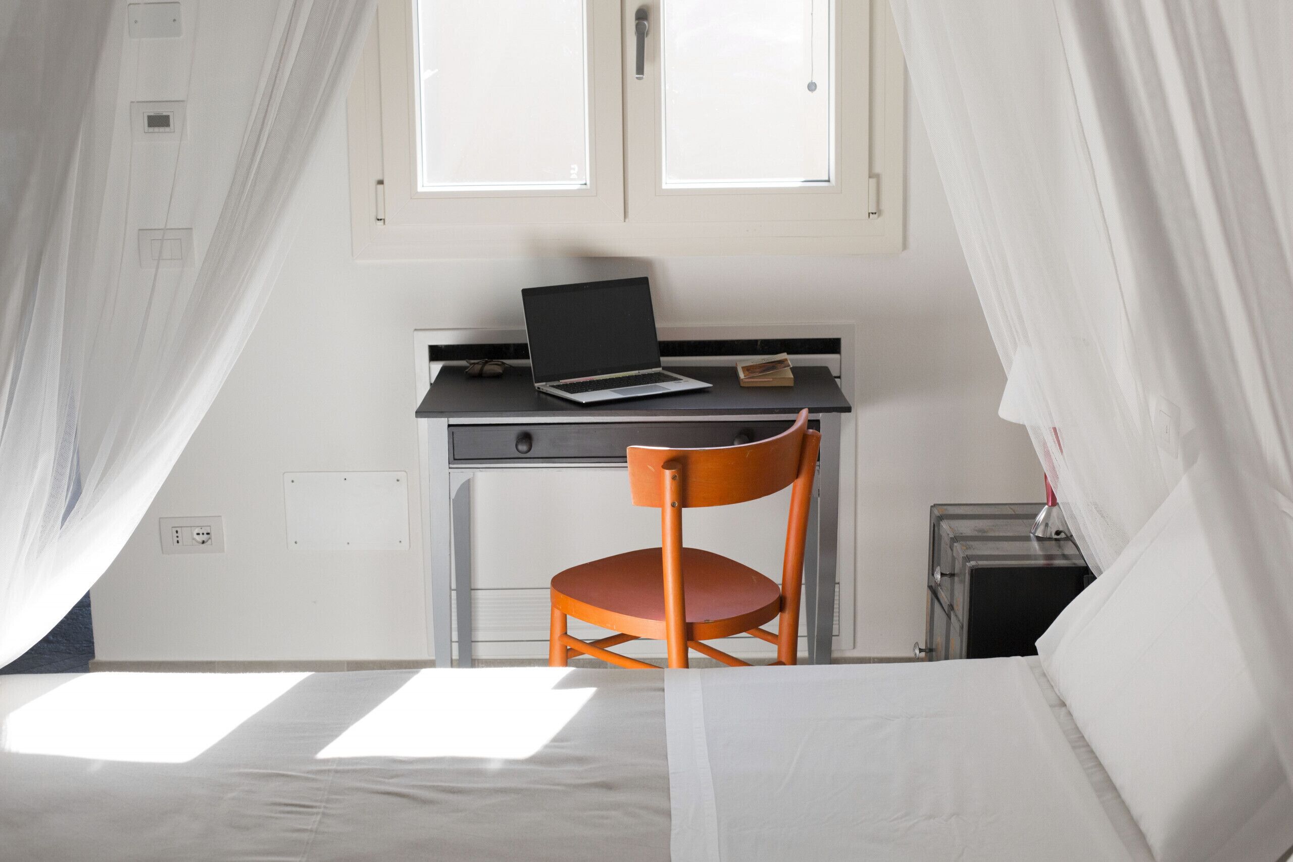 A bedroom with a bed, a desk with a laptop, a chair, and a window with curtains.