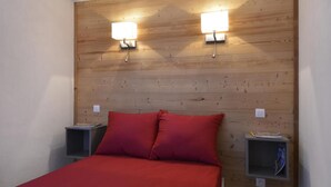A room with a wooden headboard, two bedside lamps, a bed with red bedding, and a nightstand with a book and a small item. - 100m from resort center, 3rd floor, view ski slopes, swimming pool, balcony, tv, ski locker, 35m² (La Plagne)