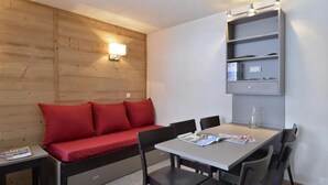 A compact room with a wooden wall, a red sofa, a dining table with chairs, and a built-in shelving unit. - 100m from resort center, 3rd floor, view ski slopes, swimming pool, balcony, tv, ski locker, 35m² (La Plagne)