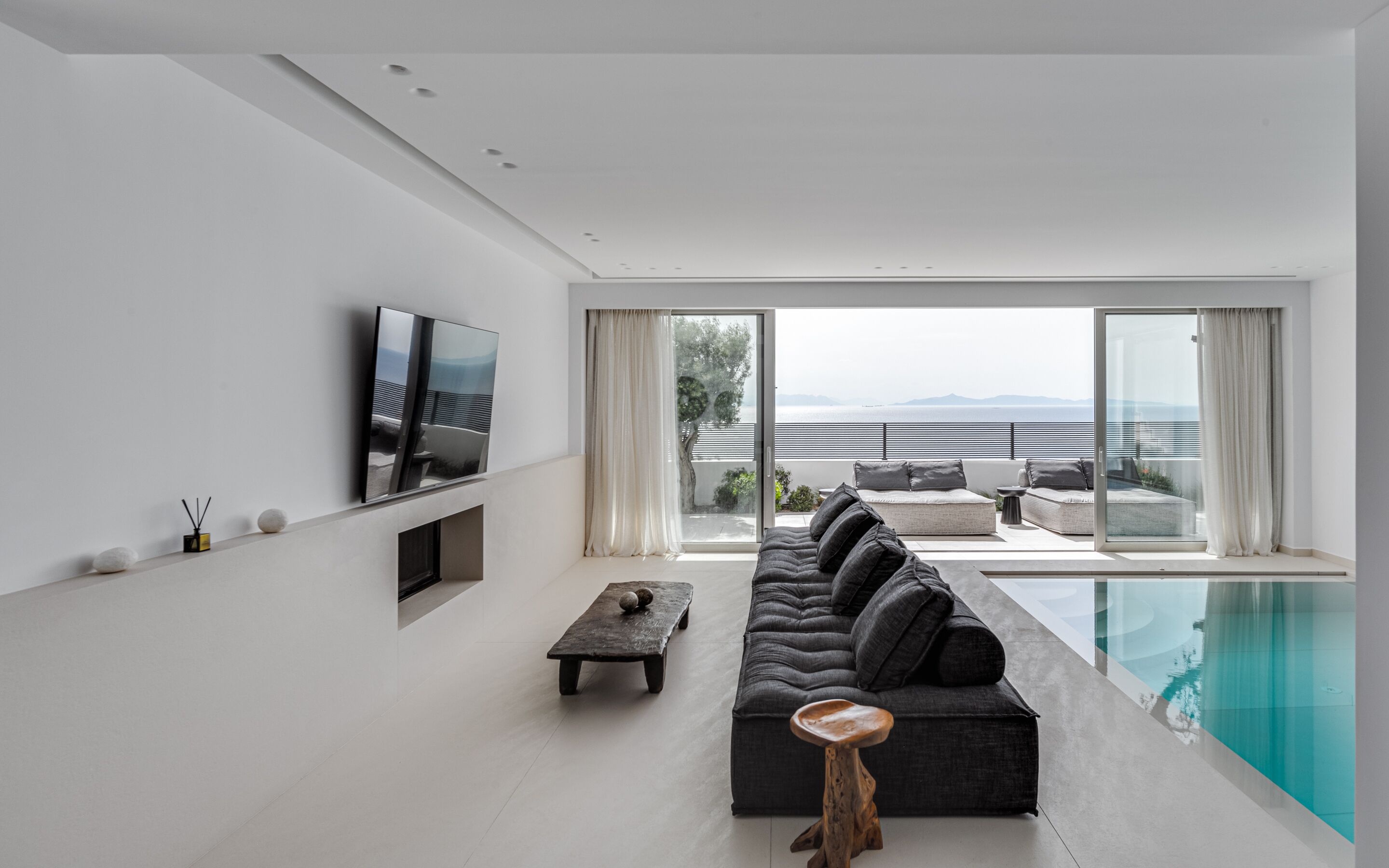 A modern living room with a large sofa, a coffee table, and a flat-screen TV mounted on the wall. There is a sliding glass door leading to an outdoor seating area with a pool.