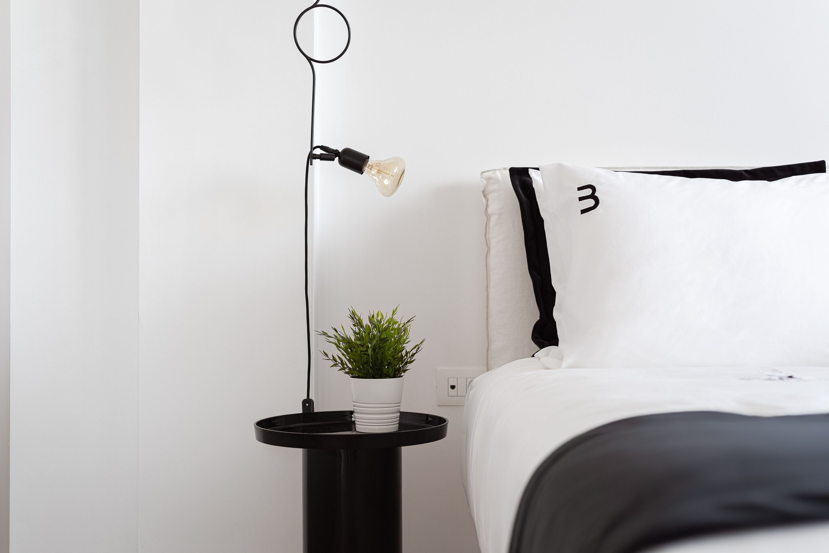 A neatly made bed with a black and white striped pillow, a small potted plant on a black side table, and a modern floor lamp with a light bulb.