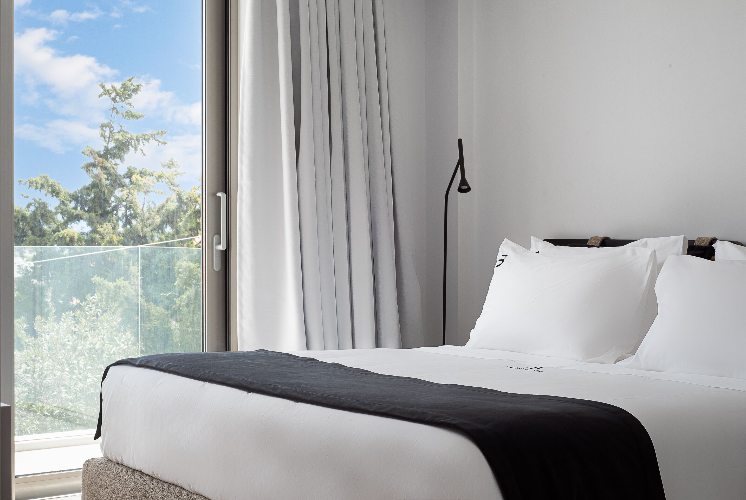 Ein modernes Hotelzimmer mit einem großen Bett, weißer Bettwäsche und Blick auf Bäume durch ein Fenster.