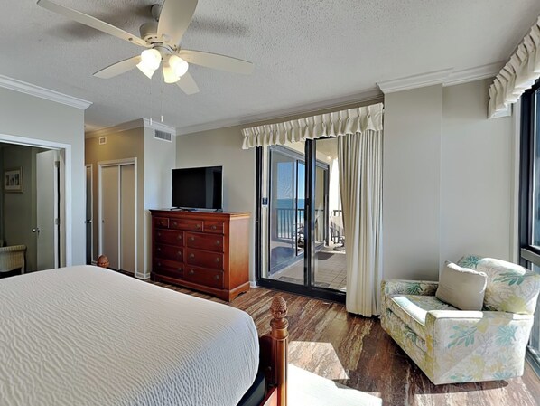 A hotel room with a bed, a dresser, a television, a balcony with a view, and a chair. - Romar House #702B | Stunning Beachfront Condo! (Orange Beach)