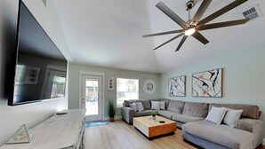 Amplia sala de estar con televisor de pantalla plana, ventilador de techo, mesa de centro de madera y sofá modular gris.