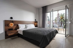 Camera d'albergo moderna con un letto grande, una testiera in legno, due comodini con lampade circolari, una porta scorrevole in vetro su un balcone e vista su palme e edifici.
