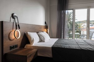 Habitación de hotel con una cama grande, cabecero de madera, dos lámparas de pared, una mesita de noche y vistas al exterior a través de un ventanal amplio.