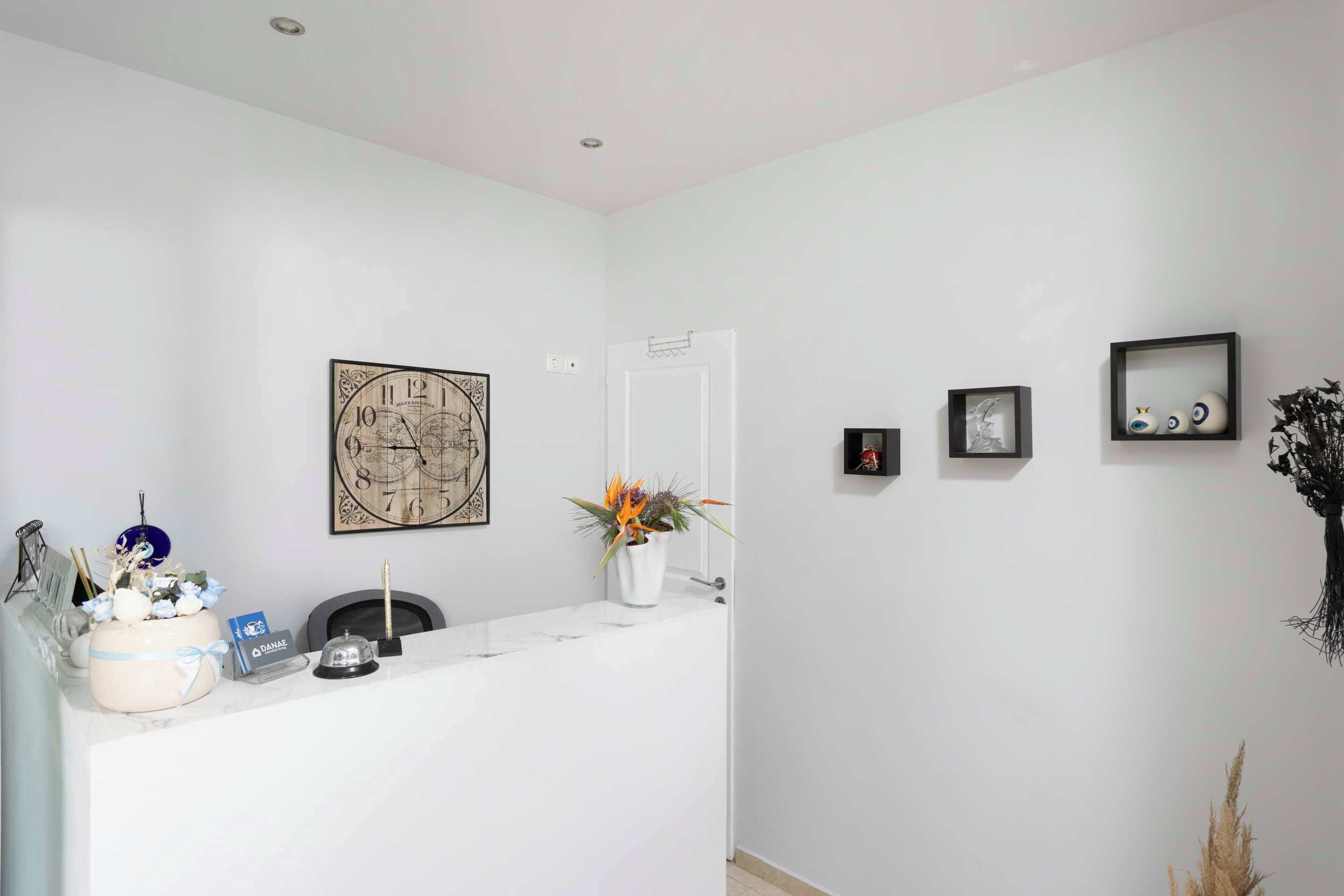 A reception area with a white reception desk, a clock, a framed artwork, a potted plant, and a small shelf with decorative items.