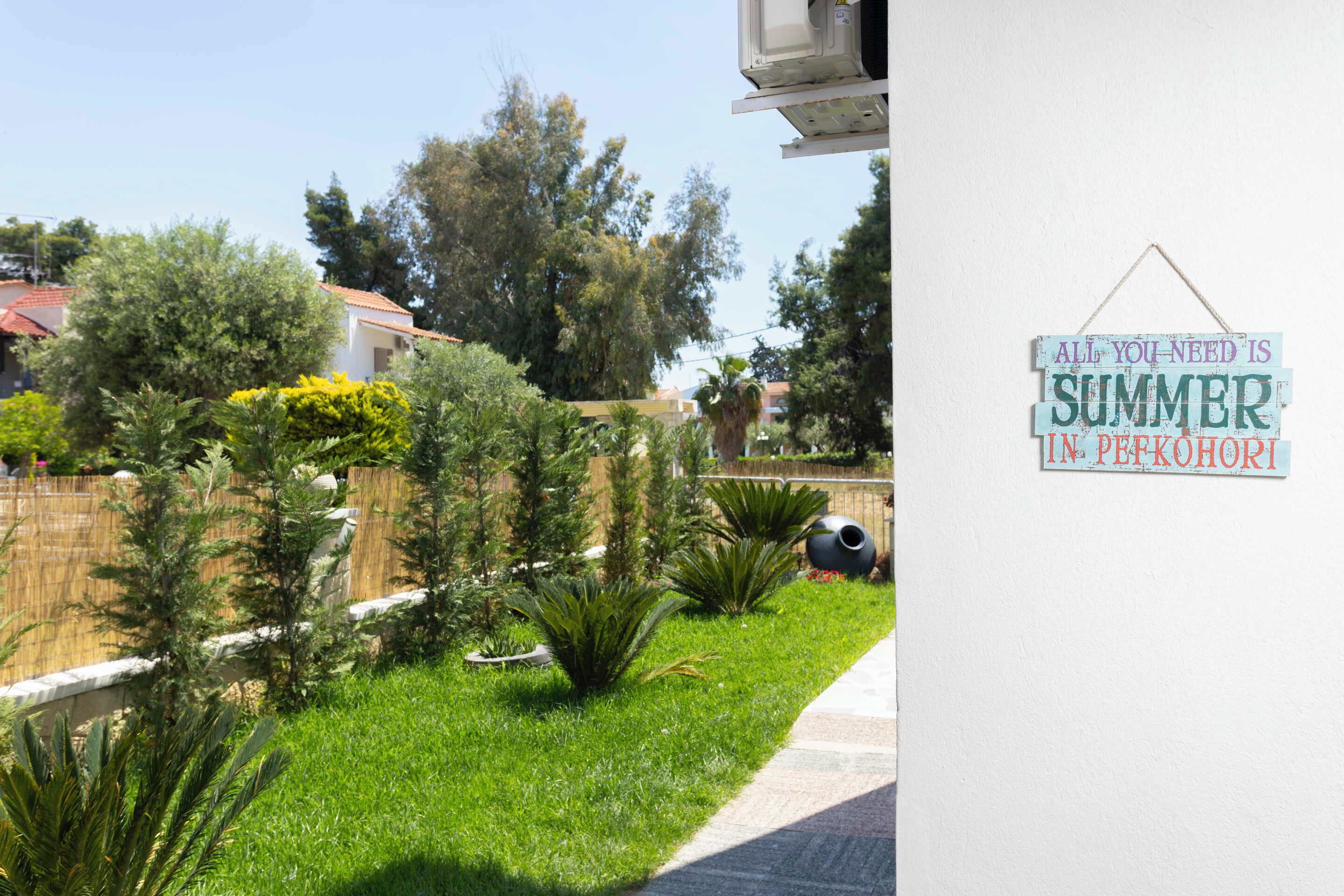A garden with various plants and a sign on a wall that reads "ALL YOU NEED IS SUMMER IN PETROHURI".