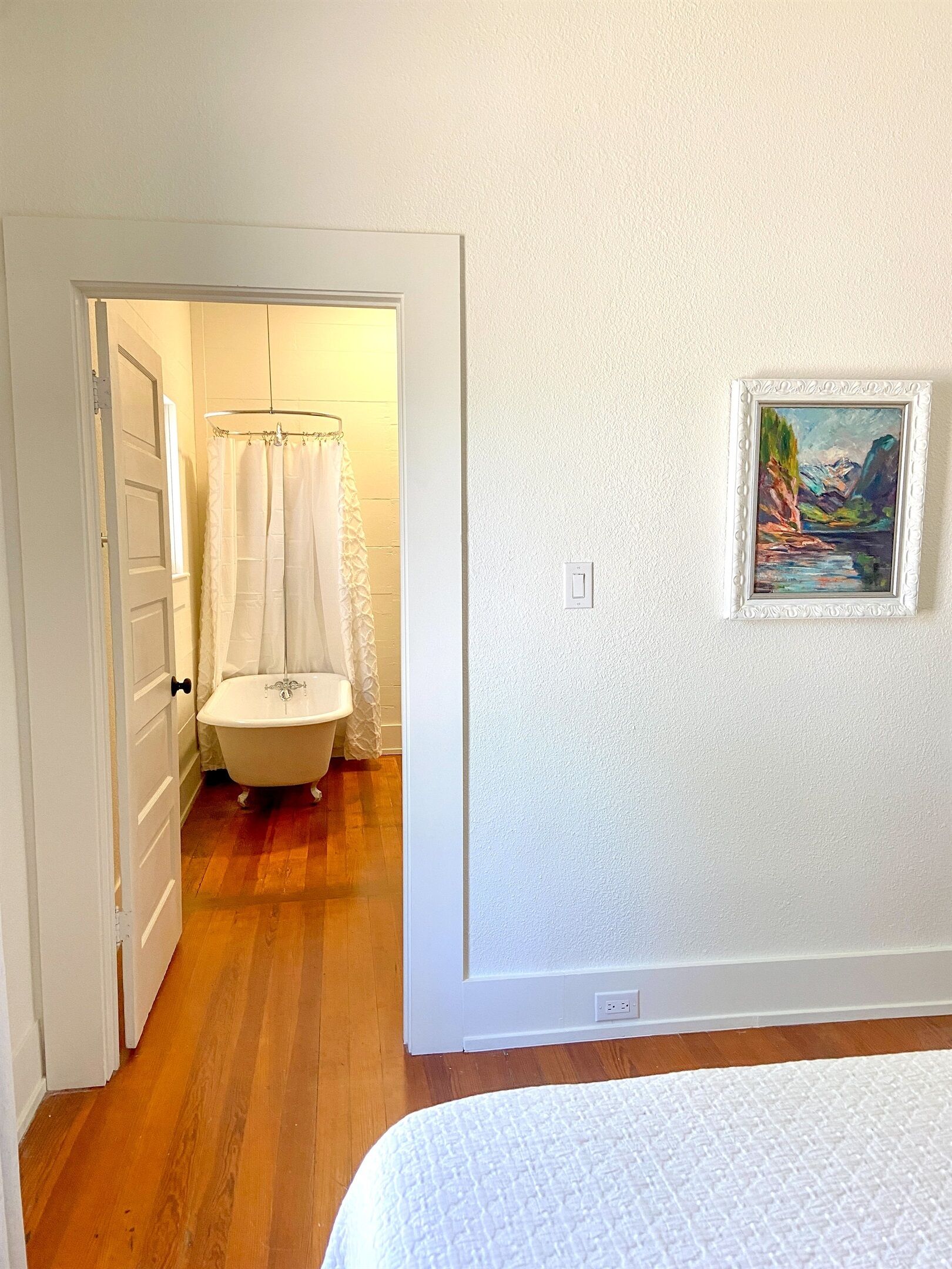 Ein Zimmer mit Holzboden, einer weißen Tür, einer weißen Badewanne und einem gerahmten Gemälde an der Wand.