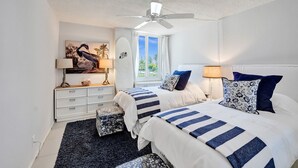Quarto com duas camas, um ventilador de teto, janela com cortinas, mesa de cabeceira com luminária e um quadro na parede.