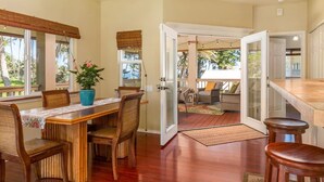 A dining area with a wooden table and chairs, a potted plant, and a view of the outdoors through large windows. - Moana Imaku - Ocean View Home w/ ADA Features (Pahoa)