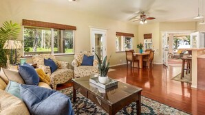A spacious living room with a wooden coffee table, two patterned armchairs, a dining area with a wooden table, and a kitchen area visible through an open door. - Moana Imaku - Ocean View Home w/ ADA Features (Pahoa)